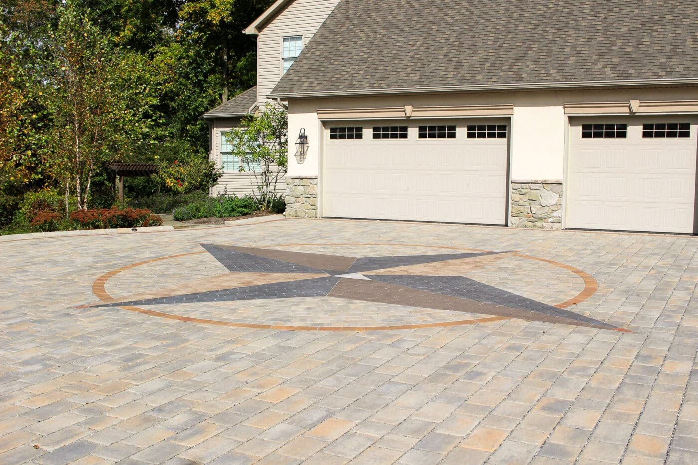 Permeable stone driveway with large compass logo in front of cream colored double garage with matching overhead doors - green trees and vegetation to the left
