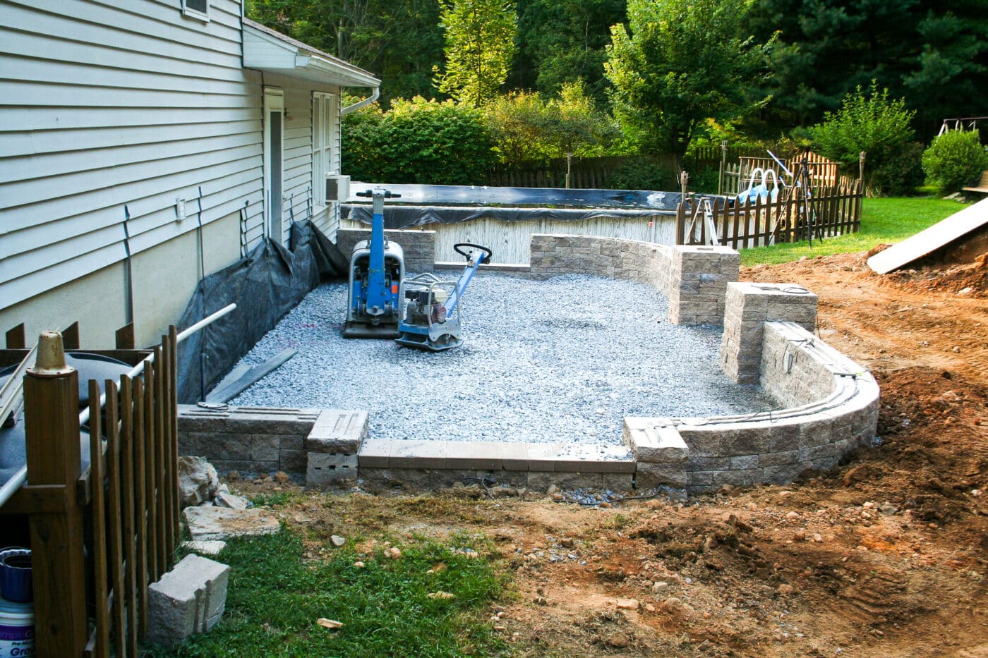 Backyard view of patio and retaining wall under construction next to pool with black cover beside lush green trees - retaining wall filled with gray stones at base and surrounded by brown dirt and a patch of grass beside off-white hose on left - blue and gray hand-operated construction equipment inside perimeter of retaining wall