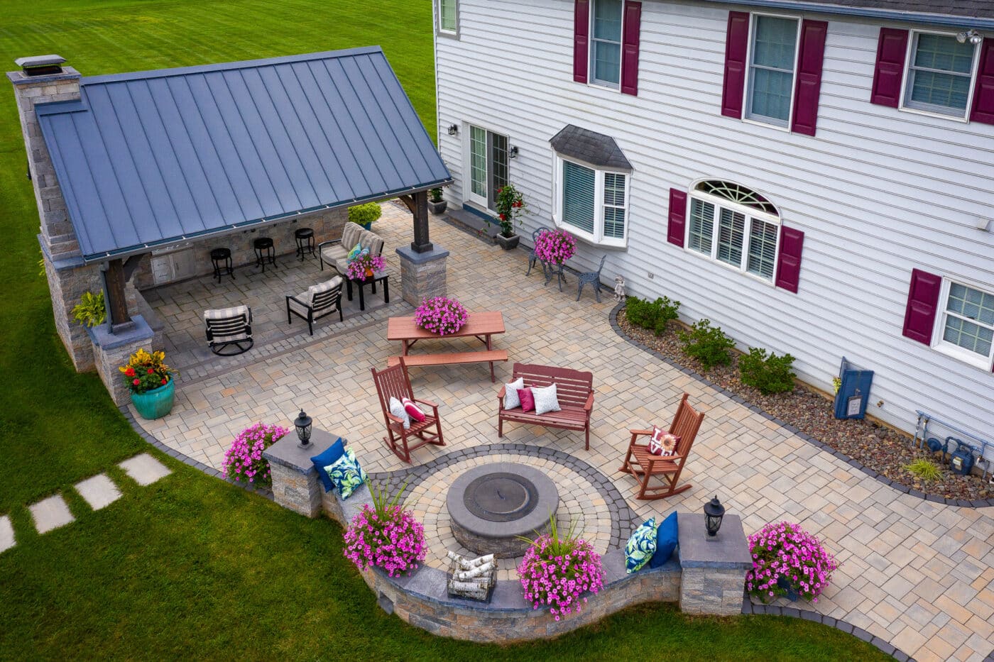 Hardscape patio with pavilion beside white two story house with six visible windows - five windows with burgundy shutters - Bar, grill and soft seating area beneath black-roofed pavillion
