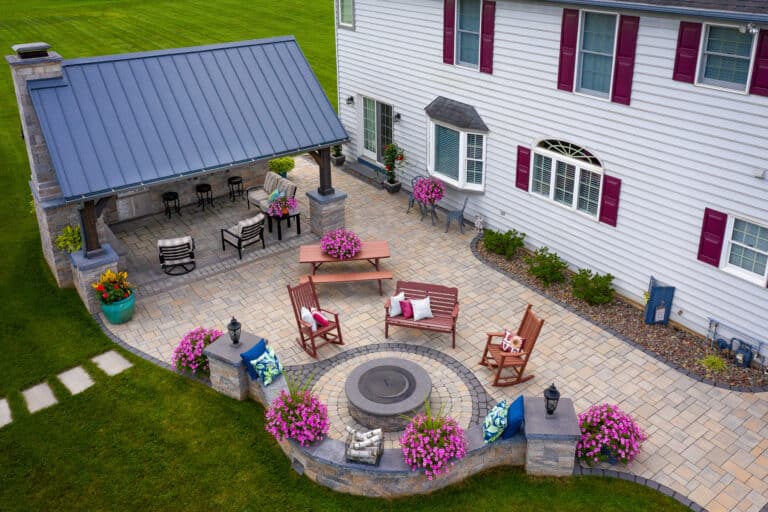Hardscape patio with pavilion beside white two story house with six visible windows - five windows with burgundy shutters - Bar, grill and soft seating area beneath black-roofed pavillion